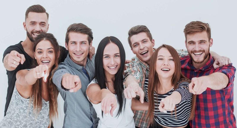Sieben junge Menschen stehen nebeneinander und zeigen lachend in die Kamera