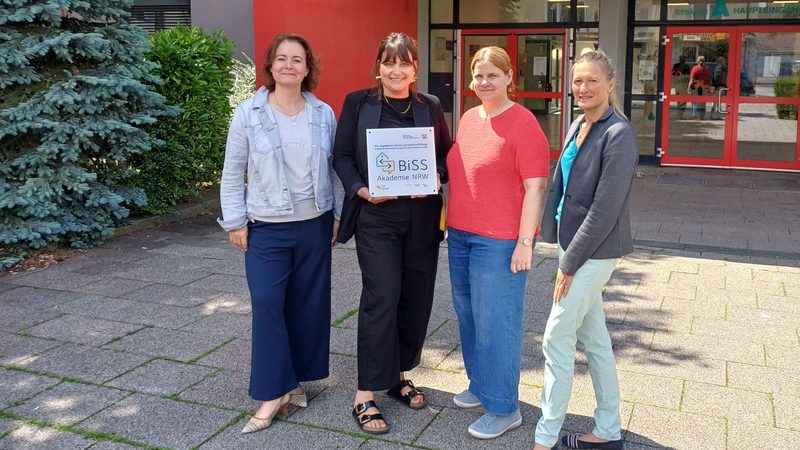 Vier Lehrerinnen stehen vor dem Schuleingang und halten ein Schild der BiSS-Akademie NRW