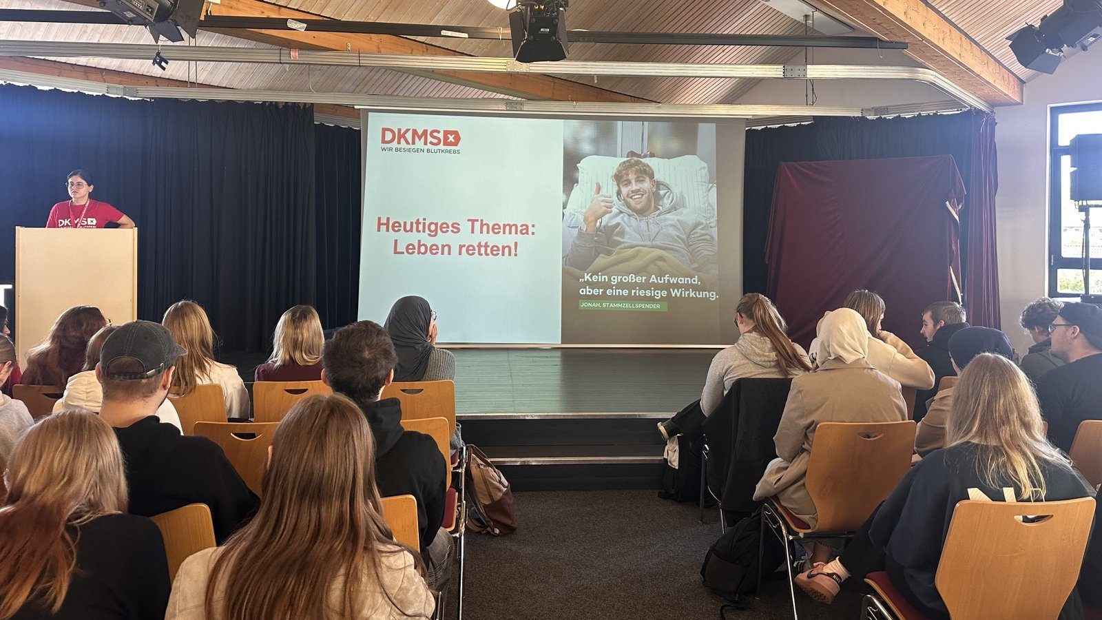 Vortrag im Forum: Eine Ehrenamtliche der DKMS steht am Rednerpult, auf der Leinwand die Folie „Heutiges Thema: Leben retten!“, im Vordergrund Schüler:innen auf Stuhlreihen