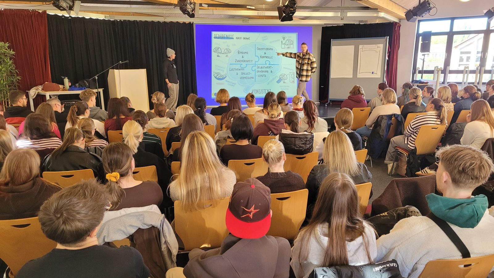 Blick von hinten über die vollen Stuhlreihen im Forum auf die Bühne, wo zwei Referierende vor einer Präsentation zu den Bedürfnissen nach Grawe stehen