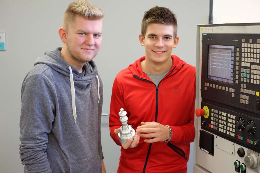 Zwei Lernende betrachten ein gedrehtes Werkstück vor einer CNC-Maschine