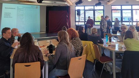 Lehrkräfte sitzen an Gruppentischen in einem hellen Saal mit Holzdecke, vorn eine Leinwand mit einer Folie der Bezirksregierung Arnsberg